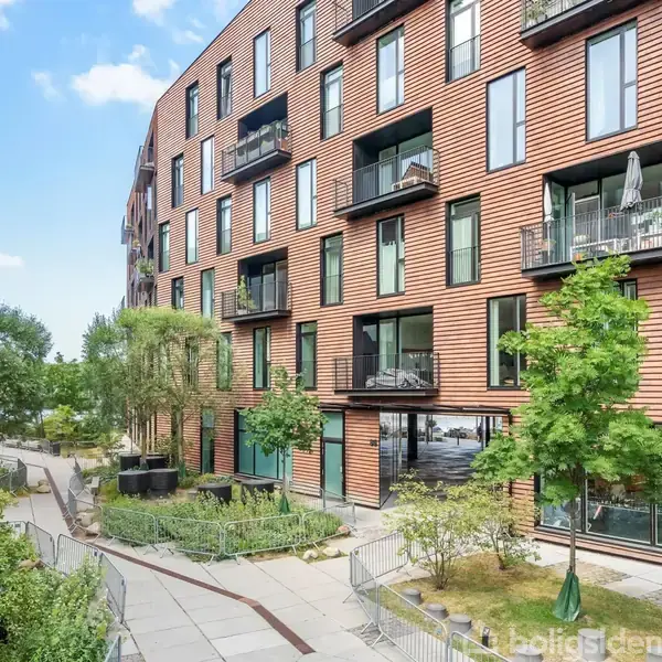 Bygning med moderne facade har altaner og store vinduer; placeret i et grønt boligområde med træer og stier.