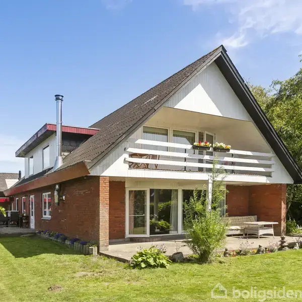 Et murstenshus med skråt tag har en balkon med rækværk; huset står i en grøn have med buske og blomster i en solrig atmosfære.