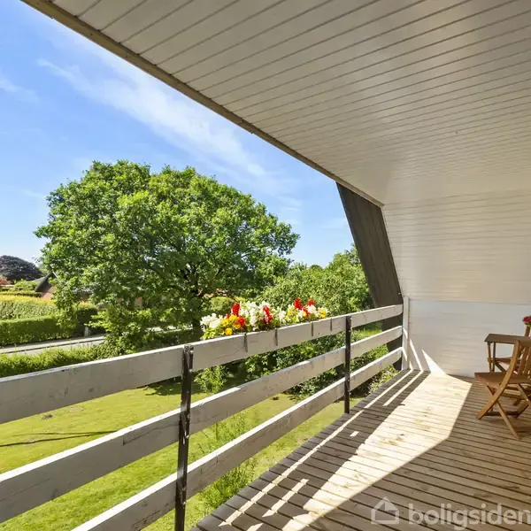 En træterrasse har et lille cafésæt med to stole og bord. Blomster pryder rækværket. I baggrunden ses en grøn have med træer, under en klar blå himmel.