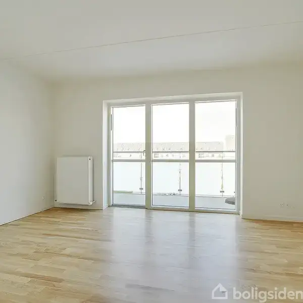 Et rummeligt, tomt værelse med lyse parketgulve. En stor glasdør åbner ud til en altan og giver udsigt til bygninger udenfor. Radiator placeret til venstre.