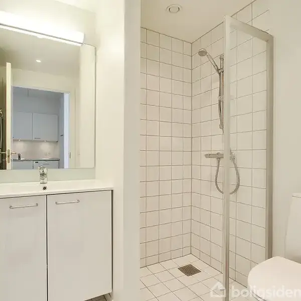 Badeværelse med hvidt flisebelagt bruseområde, toilet og skabsarrangement med spejl. Rummet er enkelt og lyst, med en åben dør, der fører til et tilstødende område.