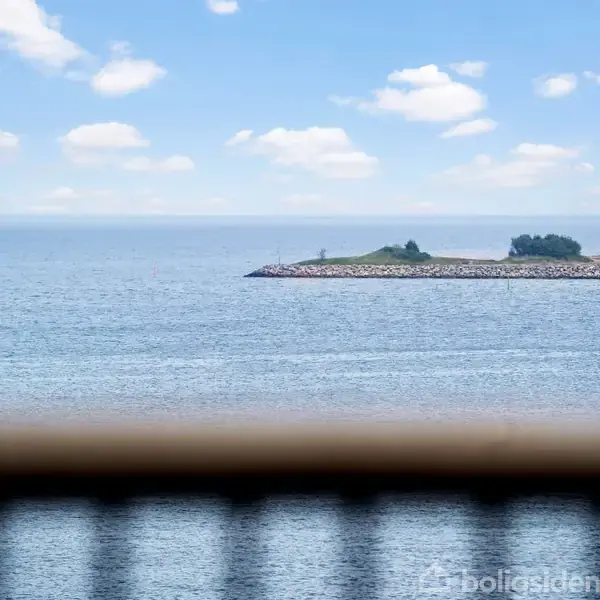 Havudsigt set gennem en træbalkon. Vandet er roligt, og i baggrunden ses en lille, grønklædt mole under en blå himmel med skyer.