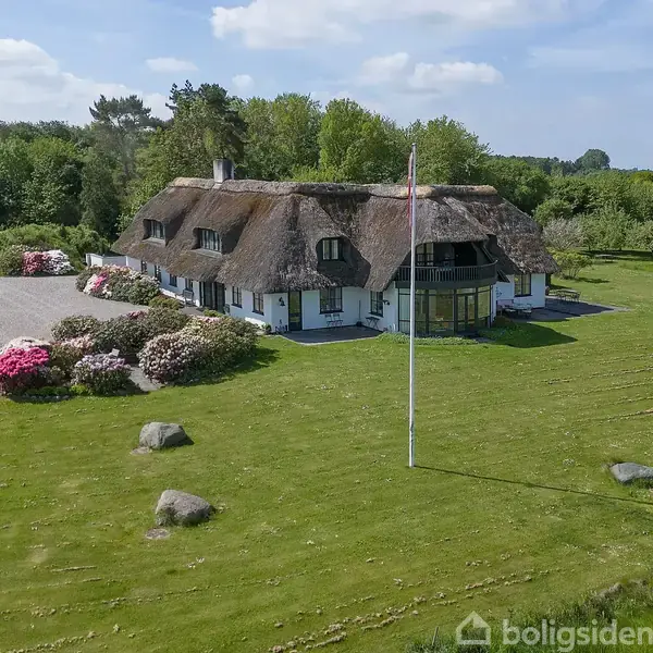 En hvidkalket stråtækt bolig står stille i en frodig have. Farverige buske pryder indkørslen, mens træer og marker omkranser stedet under en blå himmel.