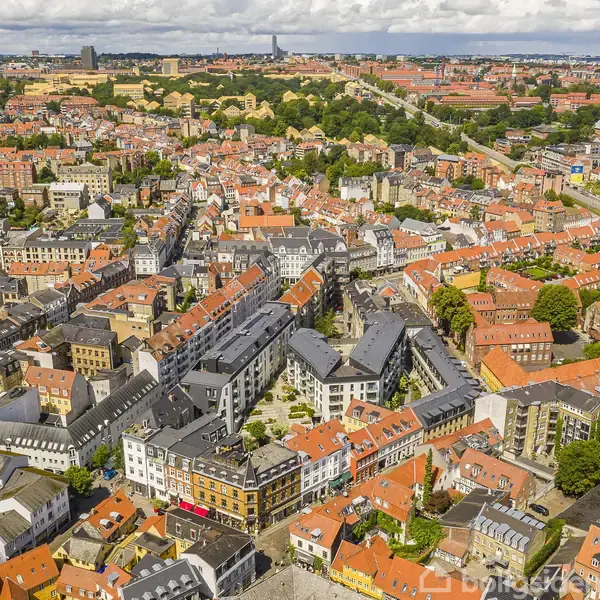 Byer med orange tage og forskellige bygninger spredt ud over et stort område. Byen ligger under en delvis skyet himmel, hvor man ser veje og grønne områder imellem husene.