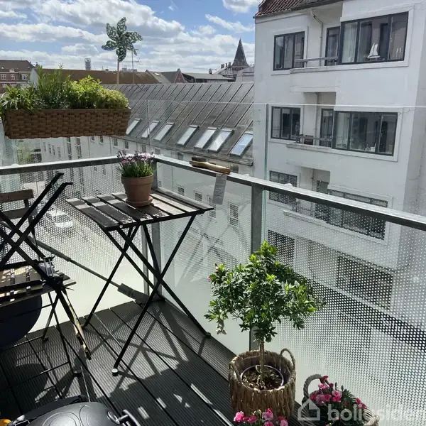 En altan med et lille bord og to stole, dekoreret med blomster og planter, omringet af høje bygninger i et moderne bymiljø.