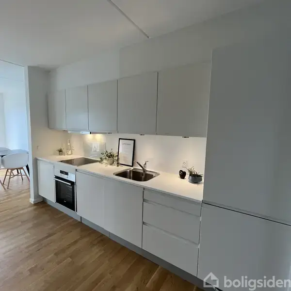 Køkken med hvide skabe og integreret ovn, vask, samt bordplade i et lyst rum. Dekorationer inkluderer en ramme på bordpladen og nogle planter. Lys træparketgulv.