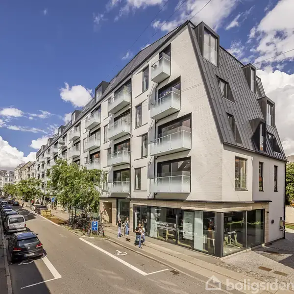 En moderne, fem-etagers bygning med store vinduer og balkoner står på en solrig gade med parkerede biler og gående mennesker. Skyer pryder en blå himmel.
