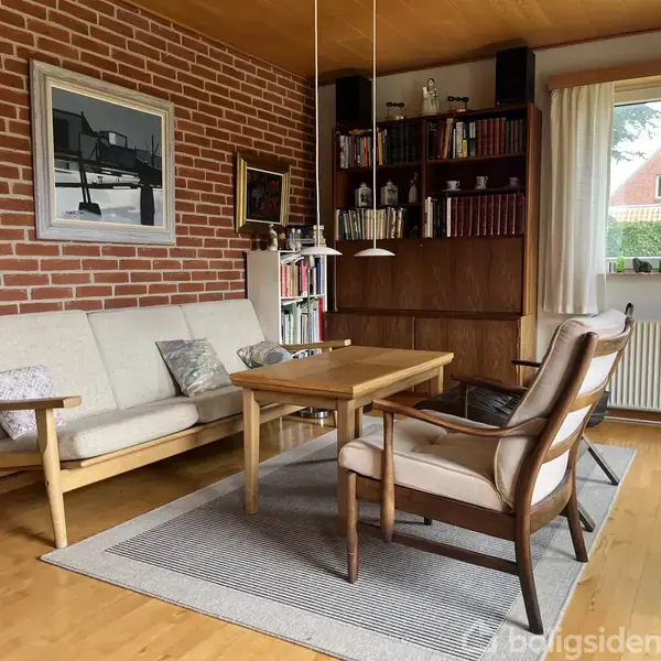 Sofa og lænestole står omkring et sofabord på et tæppe. Rummet har murstensvæg, bøger på hylder og et vindue med gardiner; billeder hænger på væggen.