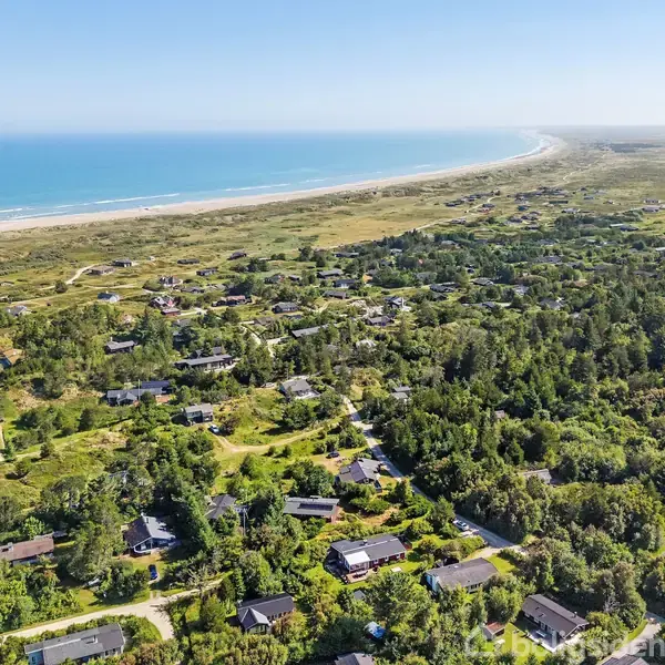 Landsbyhuse ligger spredt i en grøn, skovklædt dal ved siden af en lang, sandet kystlinje, under en klar blå himmel.
