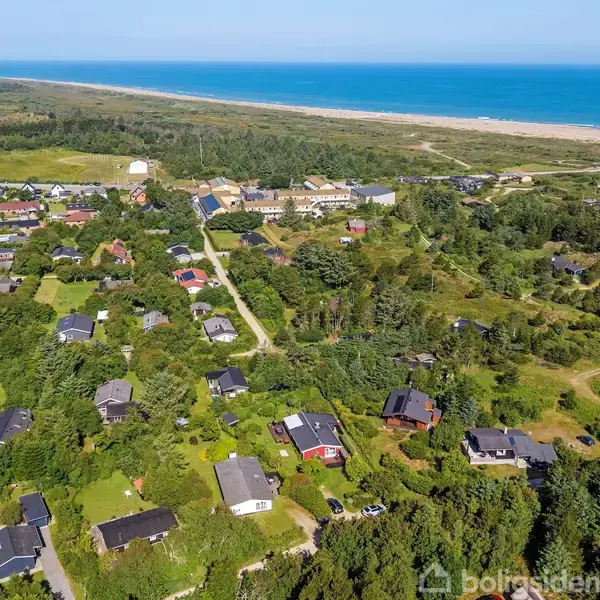 Huse ligger spredt i et grønt, frodigt område. Skove og marker omkranser landskabet. I baggrunden ses en strand og blåt hav. Området er naturrigt og landligt.