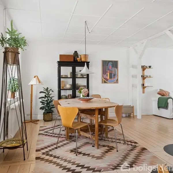Et lyst rum med spisebord og stole i midten. En brændeovn står til højre, og grønne planter pynter vindueskarmen. En reol og et maleri ses i baggrunden.