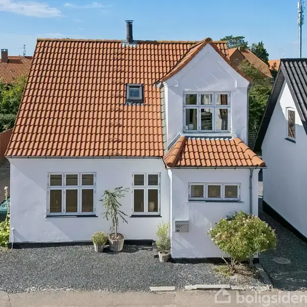 Et hvidt hus med rød tegltag står i et boligkvarter. Foran huset er der små buske og en grusbelægning. Huset har et enkeltstående skorsten og flere vinduer.