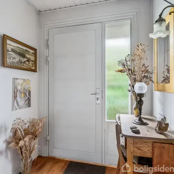 Entrédør omgivet af dekorationer; en vase med tørrede grene på et træbord, to malerier på væggen, og en velkomstskilt "Velkommen". Lyse nuancer i et hyggeligt indgangsmiljø.