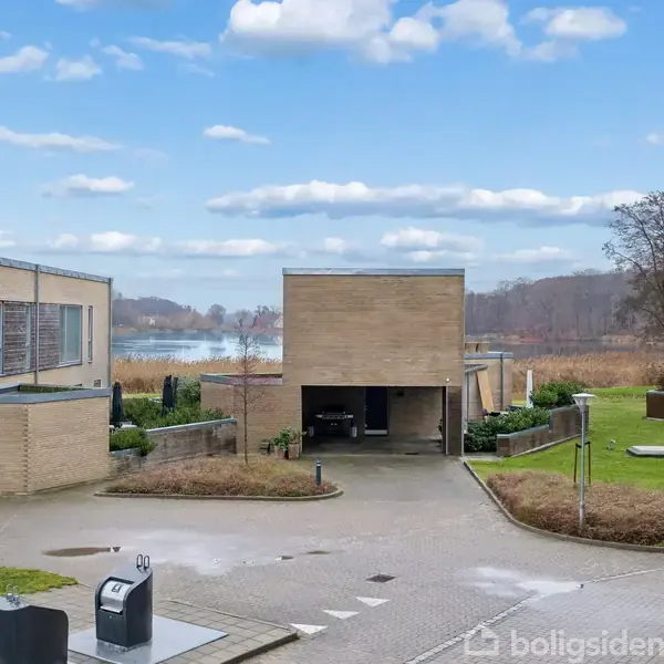 To moderne, kubistiske huse står stille i et boligområde ved en sø, omgivet af buske og træer. En parkeret bil og græs titter frem under en klar, blå himmel.