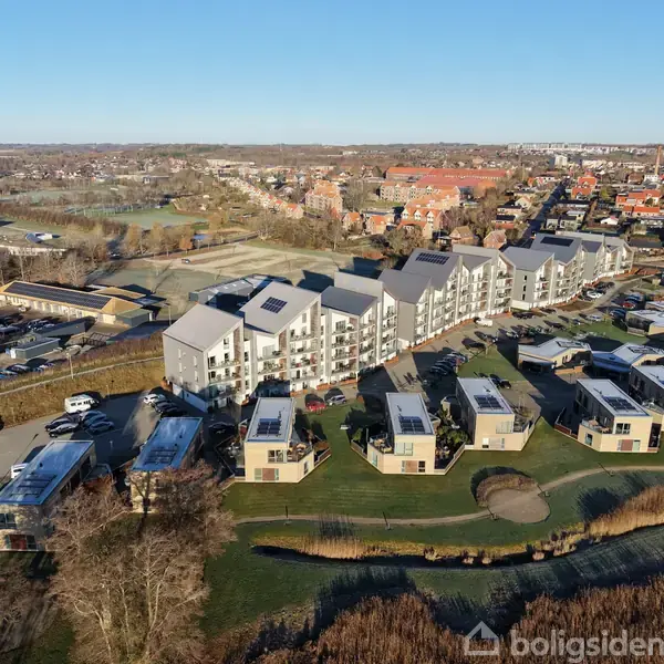 Moderne bygninger med solpaneler ligger på en bakkeskråning, omgivet af en grøn park. Biler er parkeret omkring, med en by i baggrunden under en klar himmel.