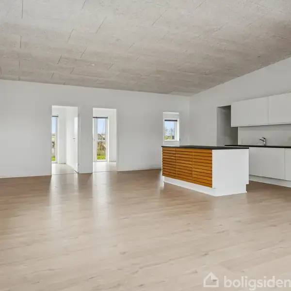 Et moderne, tomt køkken-alrum med lyse trægulve og hvidmalede vægge. Køkkenet har hvide skabe og en køkkenø med træpaneler. Rummet har flere vinduer, der giver naturligt lysindfald.
