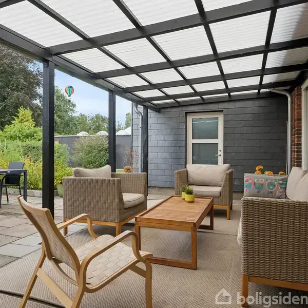 Møbleret terrasse under tag med træmøbler og hynder, placeret i en have ved et murstenshus. Der står et spisebord med stole ved siden af, mens en luftballon ses i baggrunden.