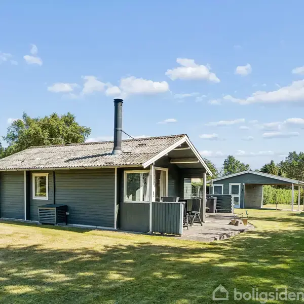 Et lille sommerhus står stille i en grøn have med spredte træer og blå himmel. En terrasse med bord og stole er placeret ved siden af huset.