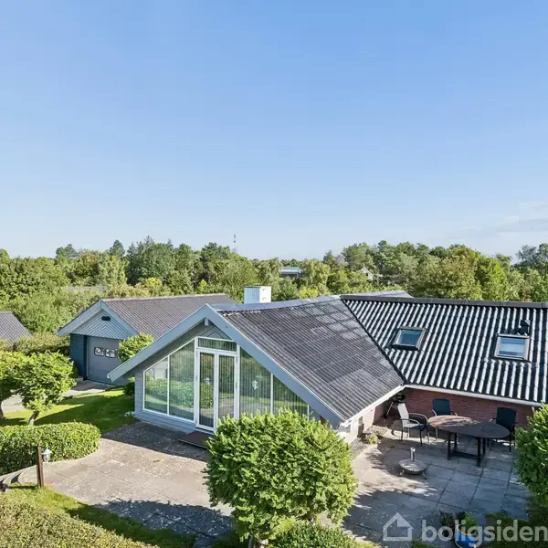 Et moderne hus med store glaspartier står i en velplejet have med flisebelagt terrasse og havemøbler. Huset har sort tag og er omgivet af frodig, grøn beplantning under en klar blå himmel.