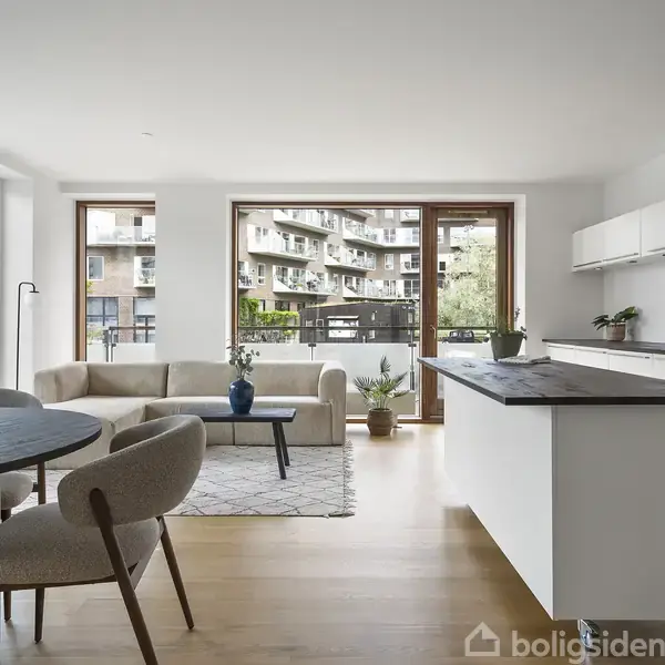 Stue/køkkenområde med sofa og rundt spisebord placeret i lyst rum. Store vinduer vender ud mod et lejlighedskompleks. Moderne indretning med planter og minimalistiske møbler.