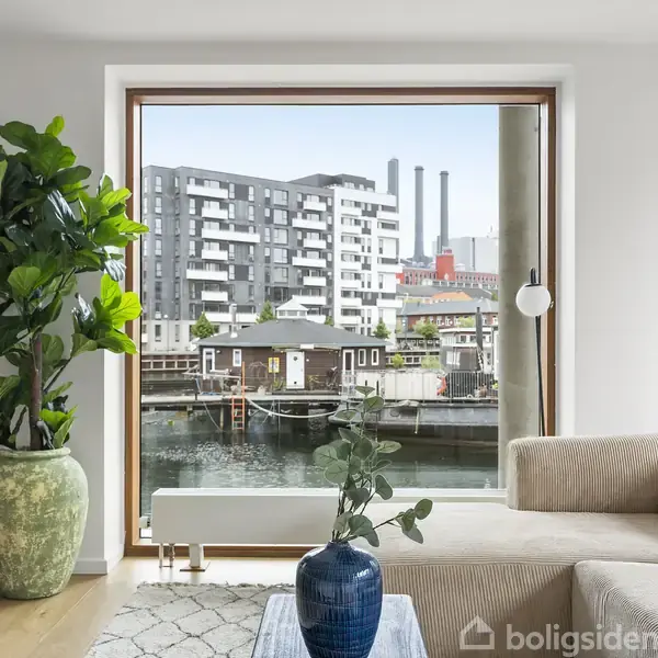 En beige sofa står ved et stort vindue med udsigt over en kanal med både og moderne bygninger. En stor grøn plante i en potte er ved siden af sofaen, og et blåt gulvvase står på et sofabord.