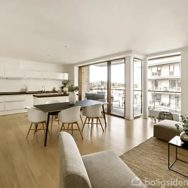 Lyst, moderne køkken-alrum med åbent design. Hvide skabe og spisebord med stole. Stue med sofabord og sofa. Store vinduer med udsigt til altan og bygninger udenfor.