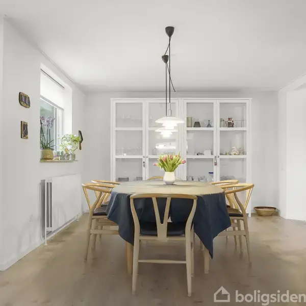 Et rundt spisebord med blå dug og vase med blomster står i midten af et lyst spiseområde. Seks træstole omkranser bordet. Indbyggede hylder og glaslåger i baggrunden.