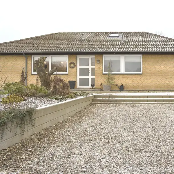 En gul murstensbungalow med fladt tag står stille i et vinterligt landskab med en grusbelagt forhave og enkelte planter tæt på en lav mur.