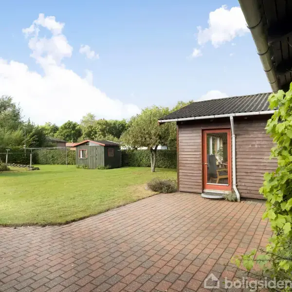 En terrasse i rød mursten ved et lille træhus med dør og vinduer, omkranset af græsplæne og buske. I baggrunden ses en grøn træhytte og en blå, skyfri himmel.