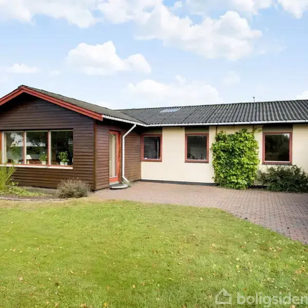 Et enplanshus med træbeklædning foran har en velplejet græsplæne og flisebelagt indkørsel. Solrig dag med få skyer danner en fredfyldt atmosfære i et boligområde.