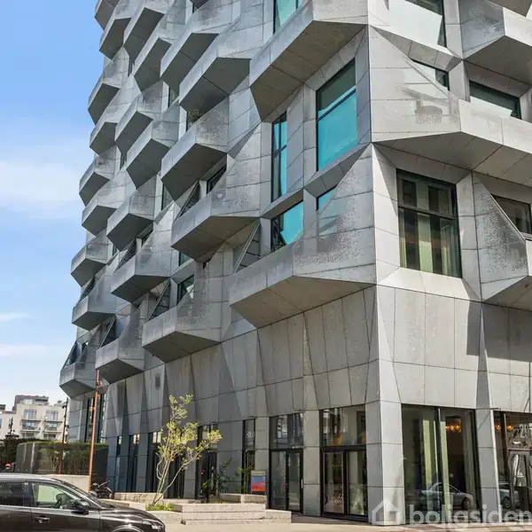 Bygning med moderne, geometrisk facade i beton og glas står i en urban kontekst. Flere udstikkende balkoner skaber et dynamisk udseende. Foran kører en sort bil langs vejen.