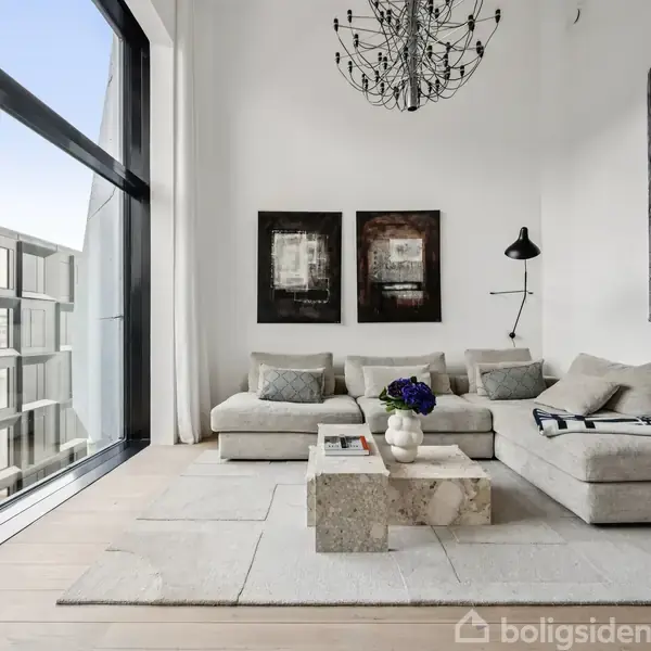 En stor, lysegrå sofa står i en moderne stue med højt til loftet. På sofabordet ligger bøger og blomster. Store vinduer slipper lys ind, og der hænger kunst på væggen.