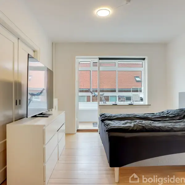 Et soveværelse indeholder en dobbeltseng med mørk sengetøj. En stor fladskærms-TV står på en kommode. Væggene er lyse, og et vindue viser hustage udenfor. På væggen hænger en whiteboard med noter.