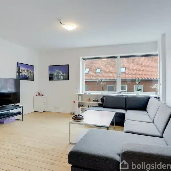 Stor, grå hjørnesofa står i en lys stue med trægulv, fladskærms-tv og billeder på væggen, mens et stort vindue viser røde hustage udenfor.