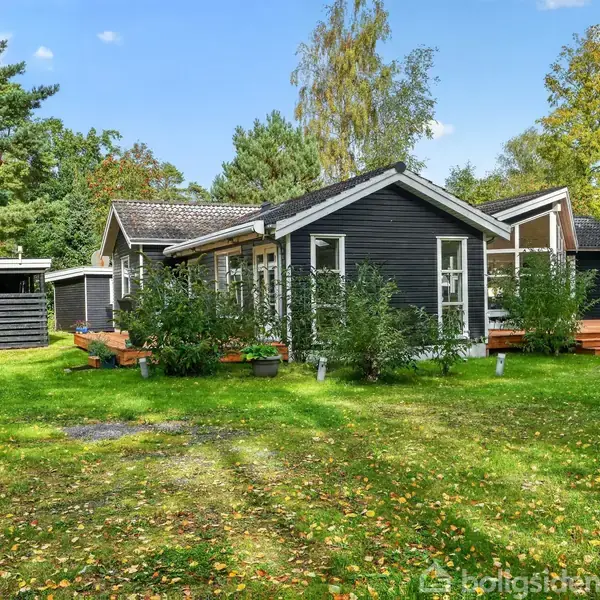 Et sort træhus står stille mellem grønne træer og græsplæne. Huset har store vinduer og en terrasse, omgivet af buske. Skyfri blå himmel og efterårsblade på plænen.