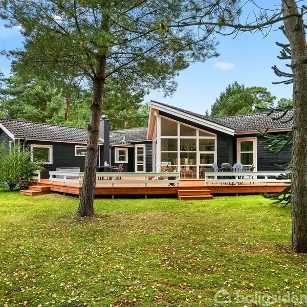 Sort træhus med store vinduer står stille i grønt skovområde. Træterrasse med møbler omkranser huset, omgivet af høje træer og græsplæne.