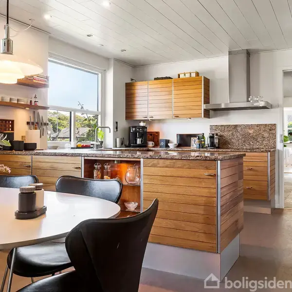 Moderne køkken med træskabe og granitbordplade, centralt placeret spisebord med blomsterdekoration; køkkenet har vindue og åbner mod en gang med trappe.