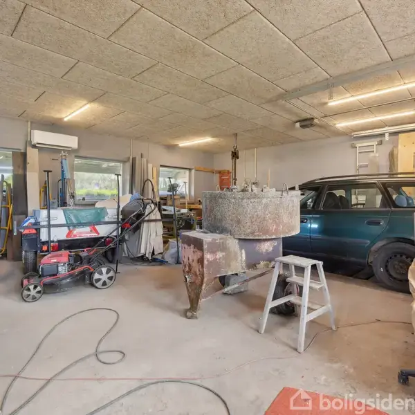 En værkstedsgarage med en blå bil til venstre. Omgivet af værktøjsudstyr, arbejdsmaskiner og diverse genstande, bord, stige. Loftslamper oplyser rummet med store vinduer bagved.