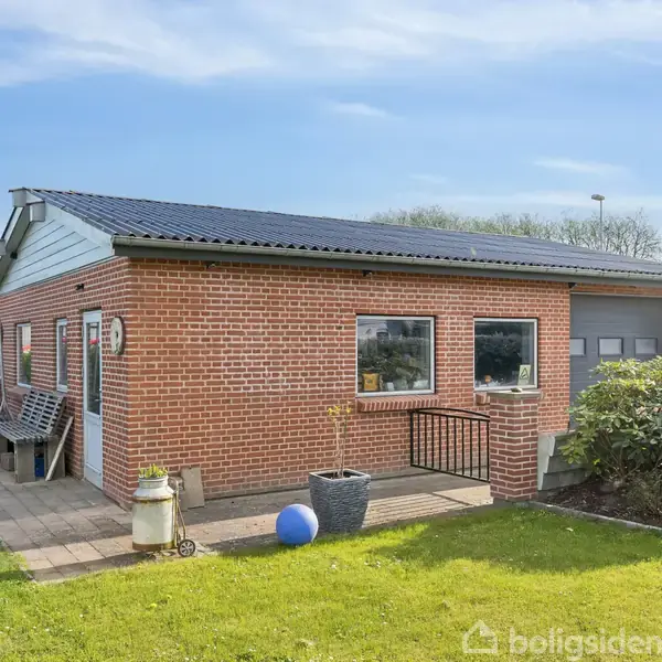 Et murstenshus med sort tag står på en grøn græsplæne. Et blåt bold er placeret ved siden af en potte på terrassen. Haven er omgivet af buske.