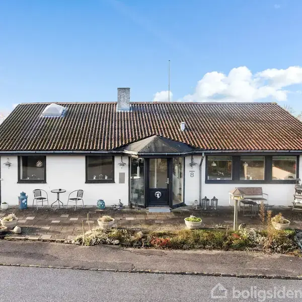 En hvid bungalow med brun tag og en indkørsel foran. Huset har en lille terrasse med møbler og potteplanter. Det ligger i et landligt område med få naboer.