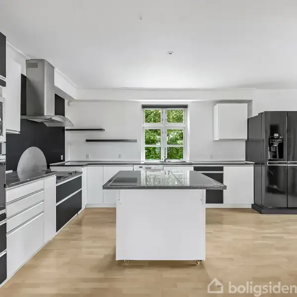 Et moderne køkken med sorte og hvide skabe, en stor køkkenø, integreret ovn og mikrobølgeovn, køleskab og emhætte, belyst af naturligt lys fra et vindue i baggrunden.