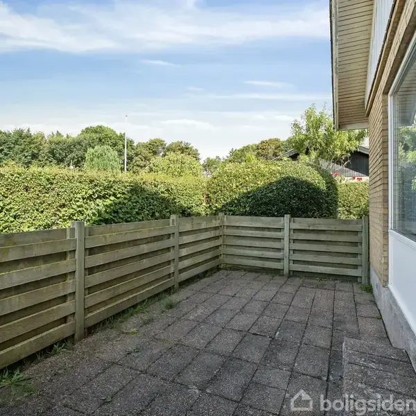 Terrasse med fliser, omkranset af et lavt træhegn. Ved siden af en gul murstensbygning med store vinduer. Baggrunden viser frodige buske og trækroner under en klar blå himmel.