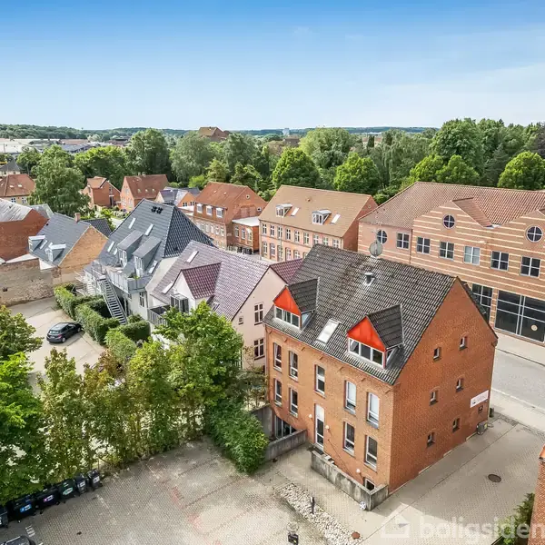 Flere huse med røde og grå tage står langs en gade omgivet af træer i et byområde. Det er en blanding af murstensbygninger og moderne huse med flere vinduer.