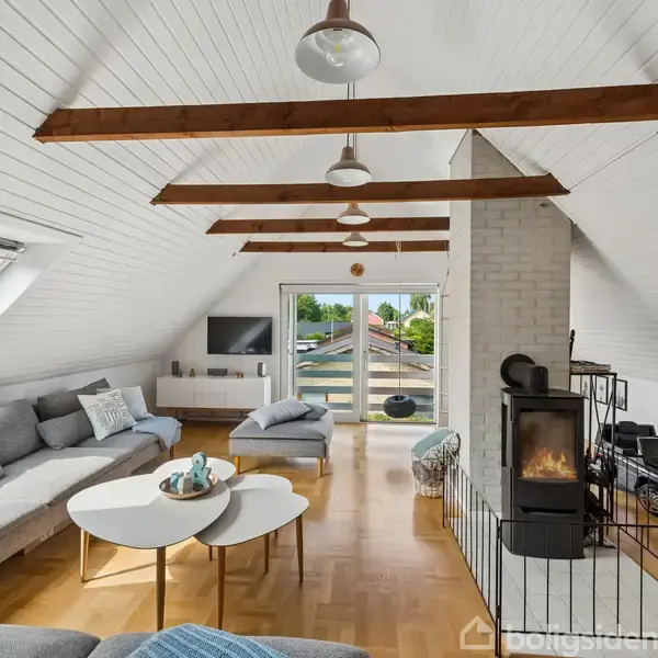 Loftstue med hvide vægge og træbjælker, stor grå sofa, to små runde borde, en brændeovn til højre, og en fransk altan med udsigt til terrasse og natur.