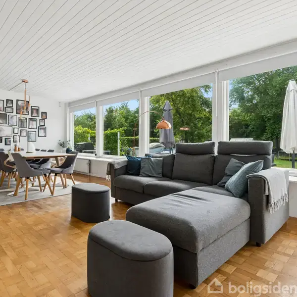 Grå sofa med puf står i en stue med trægulv og store vinduer, der viser en have. Spisebord med stole og en væg med mange billedrammer ses i baggrunden.
