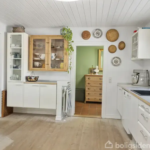 Et lyst køkken med hvidt træloft og trægulv, indeholder hvide skabe, indbygget ovn, vask og en plantede. På væggen hænger dekorative kurve og tallerkener. Baggrunden viser en dør til et andet rum med møbler.
