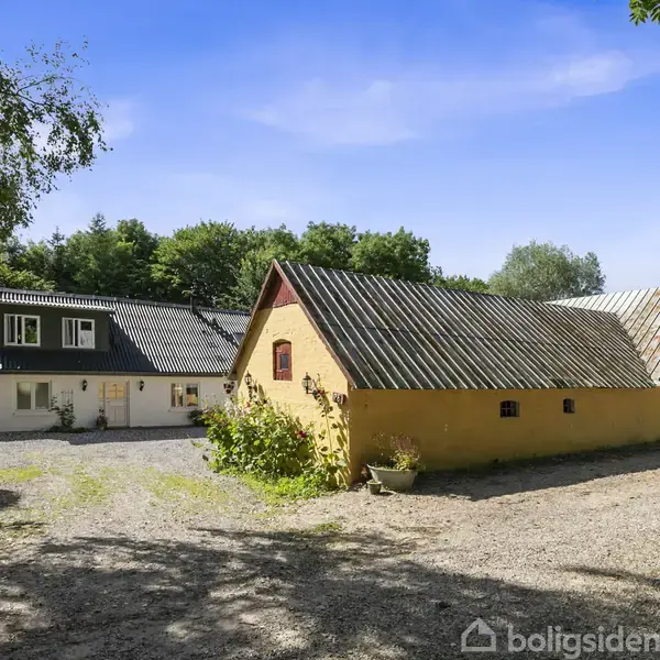 Gult hus med skråtag står på en grusvej omgivet af grønne træer. I baggrunden ses en hvid bygning med sort tag, under en klar blå himmel.