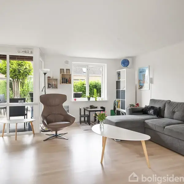 Stuen har en grå hjørnesofa og et rundt hvidt sofabord. En brun lænestol står foran en glasdør til terrassen. Vindue med udsigt til en have.