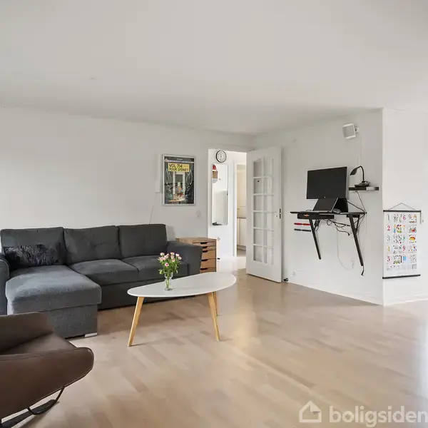 En stue med en mørkegrå hjørnesofa, der står ved en væg med to plakater. Et tv er monteret på væggen. Rummet har lyst trægulv og et hvidt sofabord.