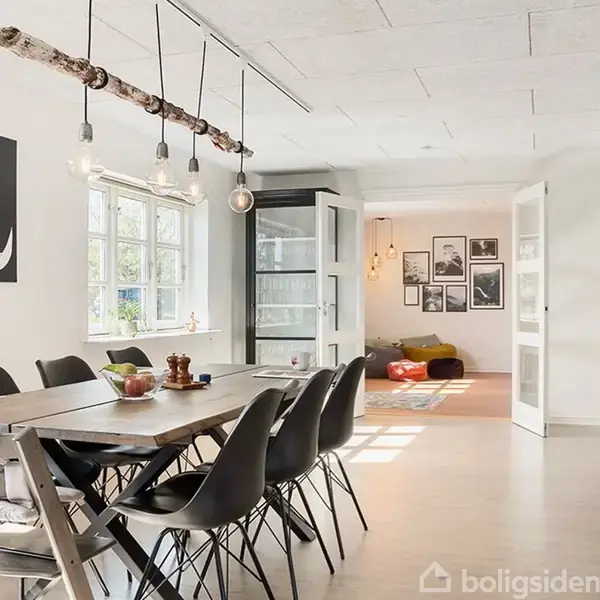 Spisebord med sorte stole står i et lyst rum ved et vindue, med kunst på væggen bagved. I baggrunden kan ses en hyggelig stue med sofaer og billeder.